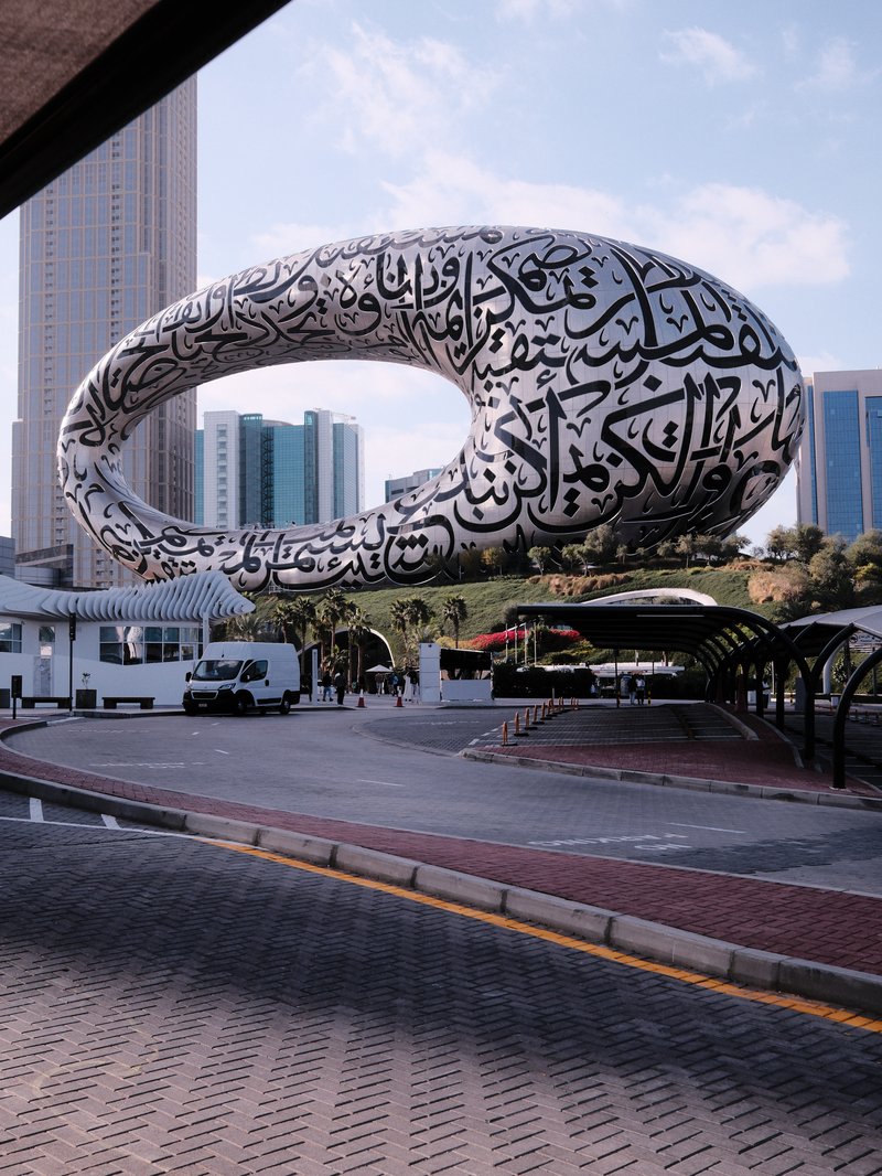 A large, modern sculpture with Arabic calligraphy in an urban setting, surrounded by buildings and vehicles.