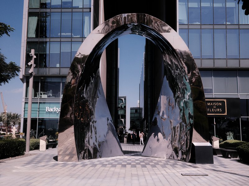 A large, reflective arch sculpture in an urban setting with modern buildings in the background.