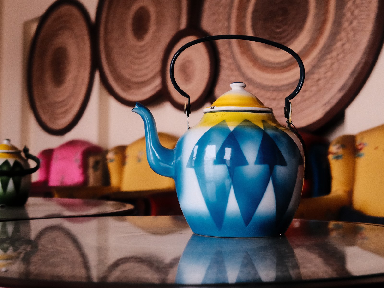 A colorful blue teapot with geometric patterns on a table, surrounded by vibrant chairs and woven wall decor.
