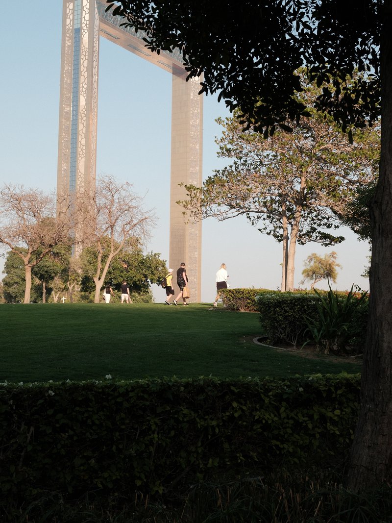 Two people walking in a park near a large architectural structure under a clear sky.