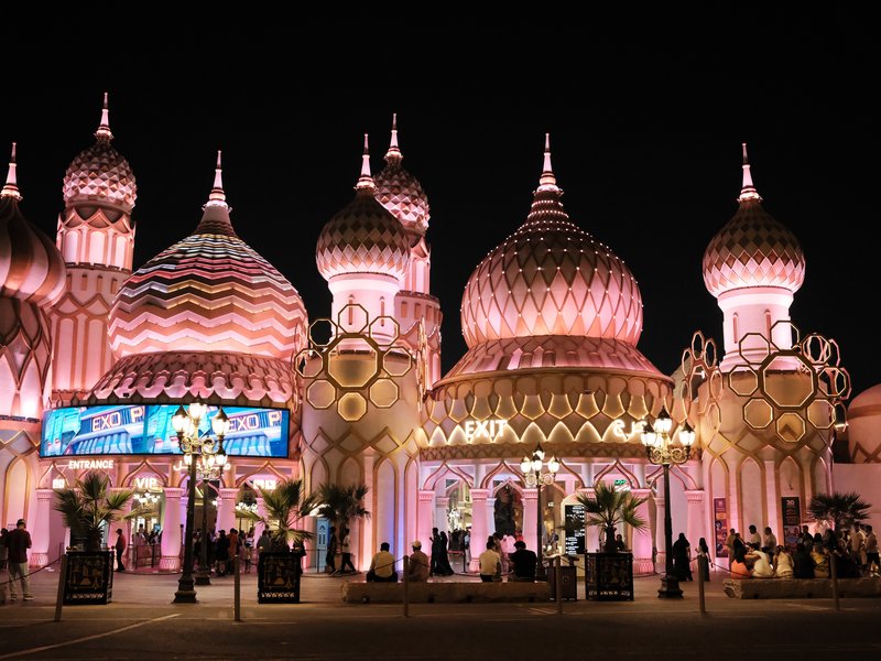 Colorful illuminated buildings with domes and spires at night, surrounded by palm trees and people.
