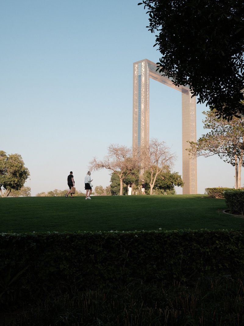 Two people walking on a grassy area with a large architectural structure in the background under a clear sky.
