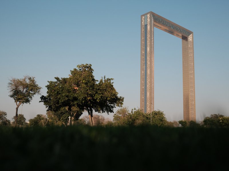 A large modern structure resembling a frame stands against a clear blue sky, with trees in the foreground.