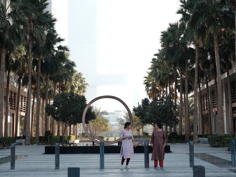 Two people standing in a palm-lined urban plaza with a circular sculpture in the background.