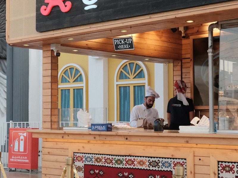 Two chefs working behind a wooden food stall with colorful windows in the background.