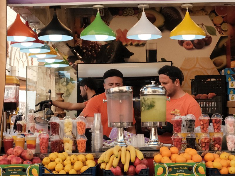 A vibrant fruit and juice stall with two vendors in orange shirts, surrounded by colorful fruit displays.