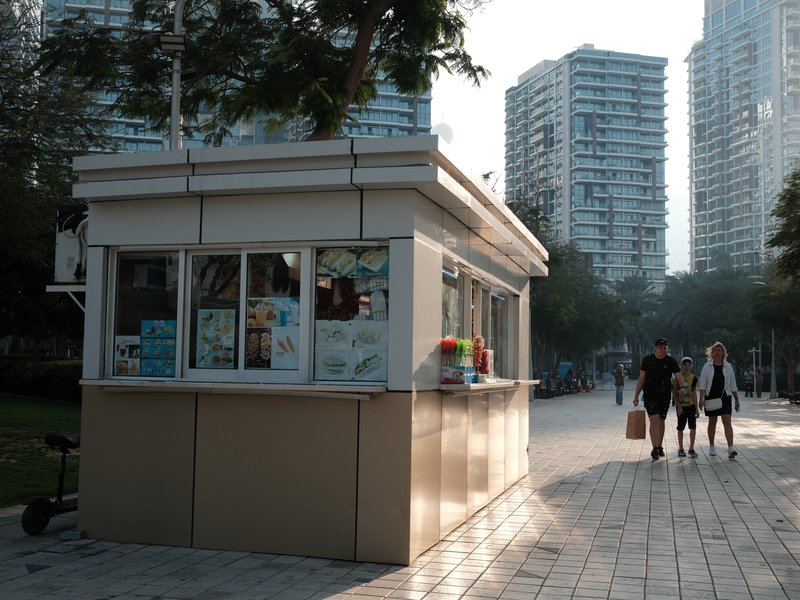 A small kiosk with large windows on a walkway, surrounded by tall buildings and people walking.