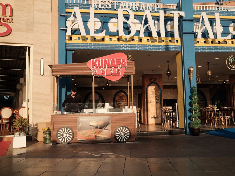 A food cart named 'Kunafa' in front of a restaurant with colorful signage and outdoor seating.