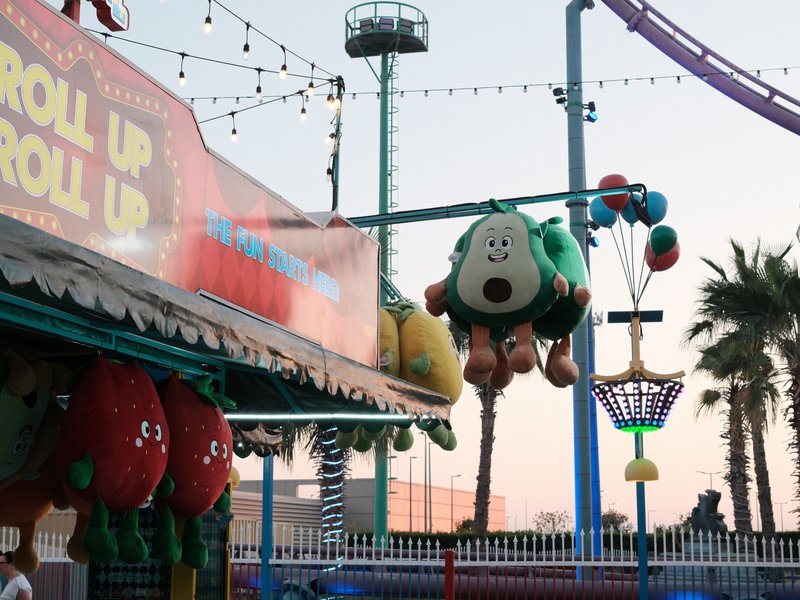Colorful carnival scene with plush toys hanging and a sunset sky in the background.
