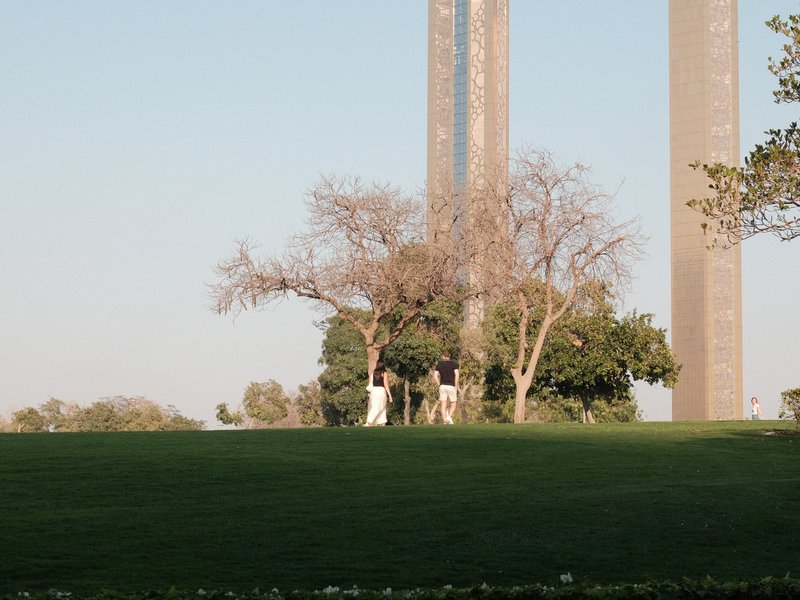 A group of people standing under trees with tall structures in the background on a clear day.