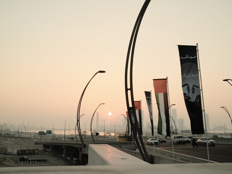 Sunset over a waterfront with tall banners and streetlights silhouetted against the sky.