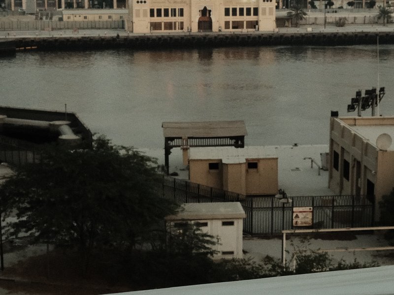 A view of a riverbank with buildings and trees, reflecting in the water under soft lighting.