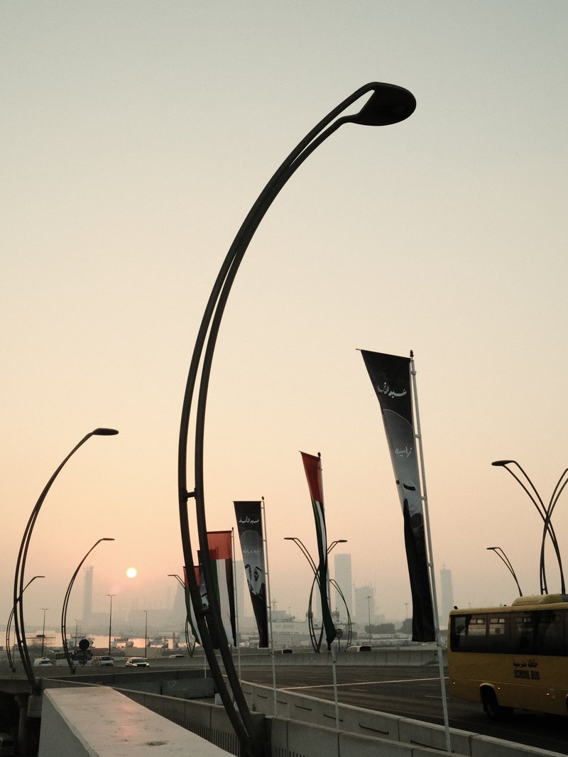 Curved street lamps and banners silhouetted against a sunset over a waterfront.