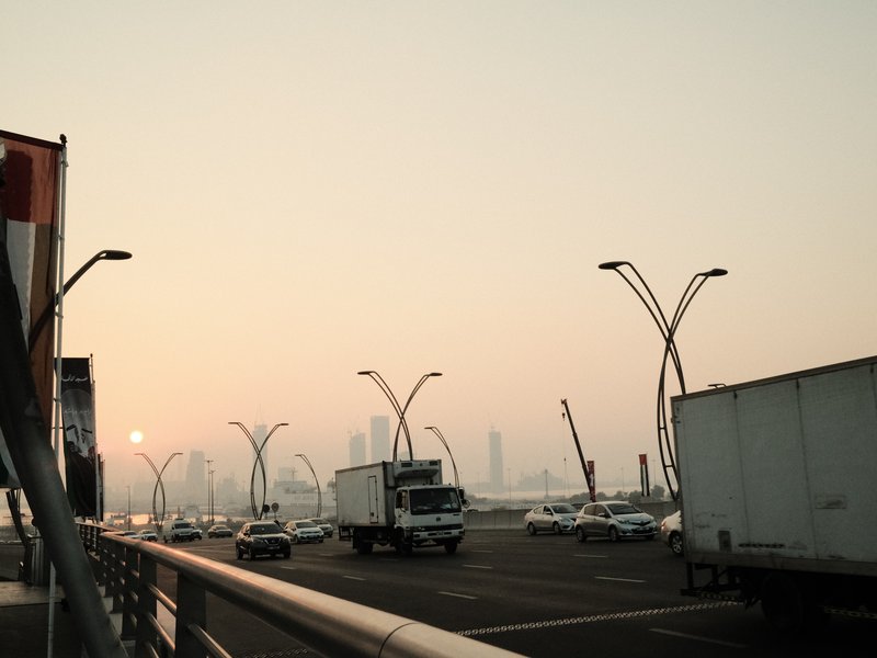 A hazy urban scene with trucks and cars on a road, skyscrapers in the background during sunset.