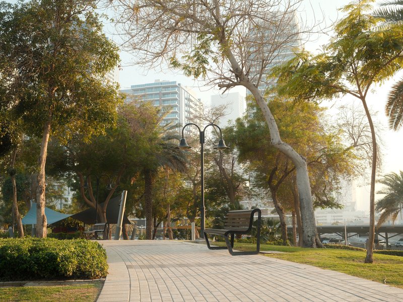 A serene park scene with a bench along a paved path, surrounded by trees and modern buildings in the background.