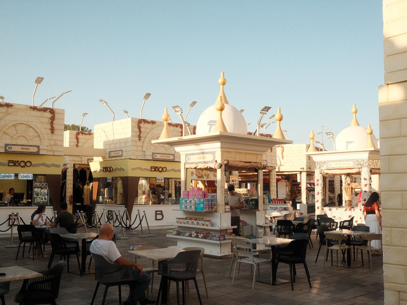 Outdoor market scene with colorful stalls and people seated at tables under a clear sky.
