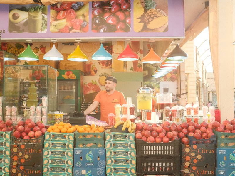A fruit vendor stands behind a colorful display of fruits in a market, with vibrant hanging lights above.