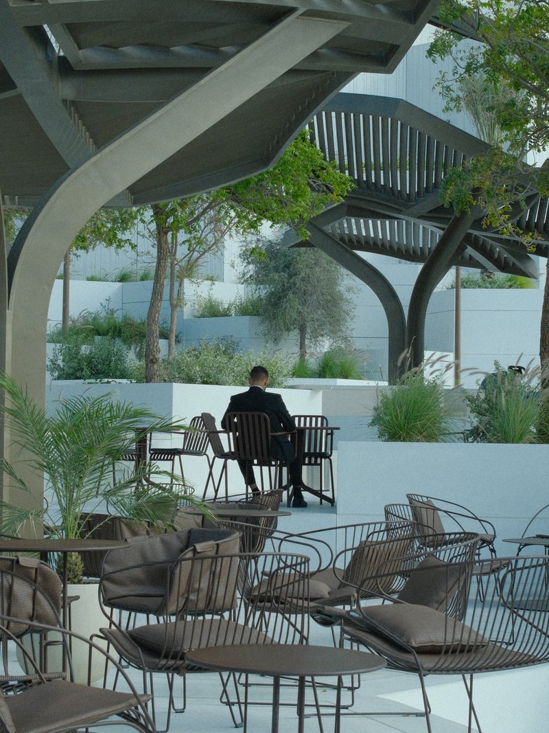 A solitary figure seated at a modern outdoor café surrounded by empty chairs and greenery.