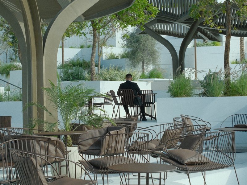 A person sitting alone at a table in a modern outdoor café surrounded by greenery.