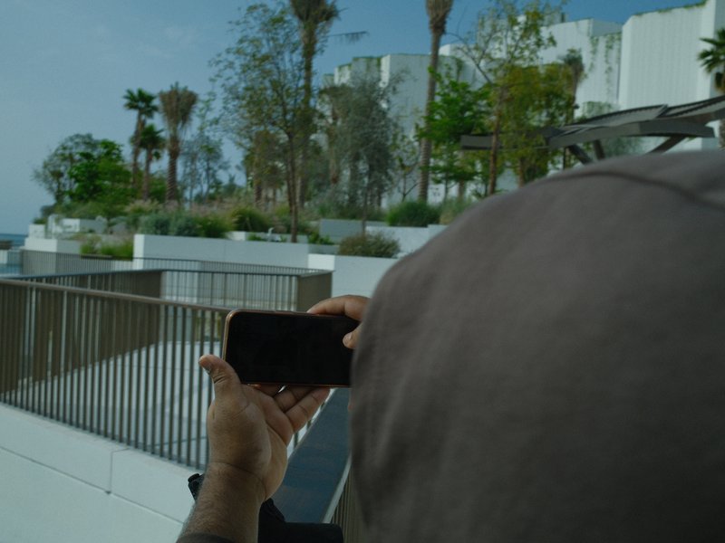 A person holding a smartphone in an outdoor setting with palm trees and modern architecture in the background.