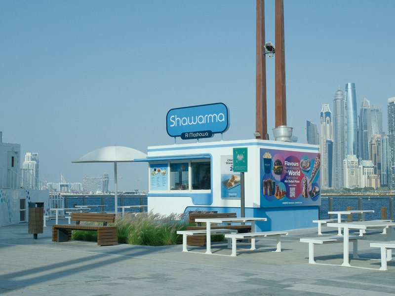 A bright blue food kiosk named 'Shawarma' near a waterfront with city skyline in the background.