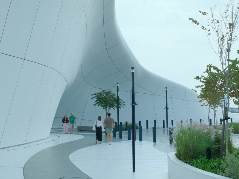 Modern architectural structure with curved lines, people walking along a path, greenery nearby.