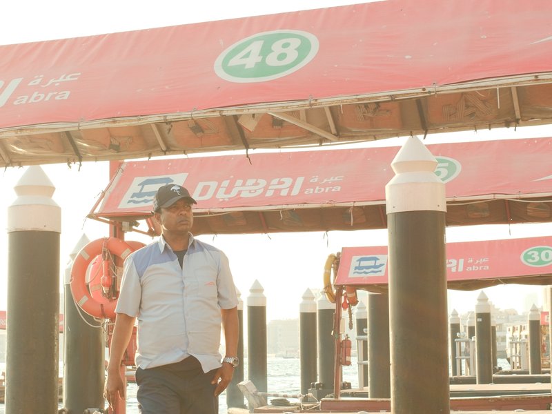 A man walking under a dock with red canopies and boat numbers, sunlight creating a warm atmosphere.