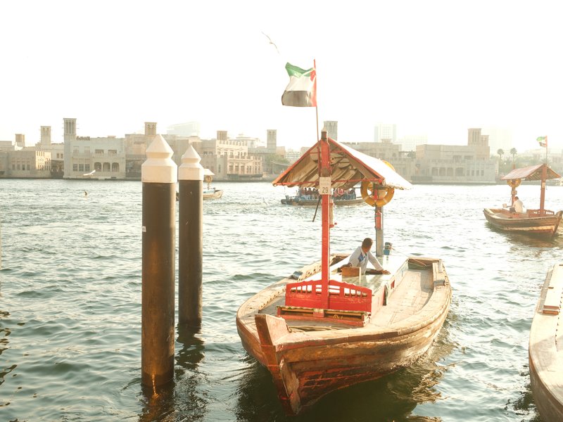 Traditional wooden boat docked near waterfront with city buildings in the background under bright sunlight.