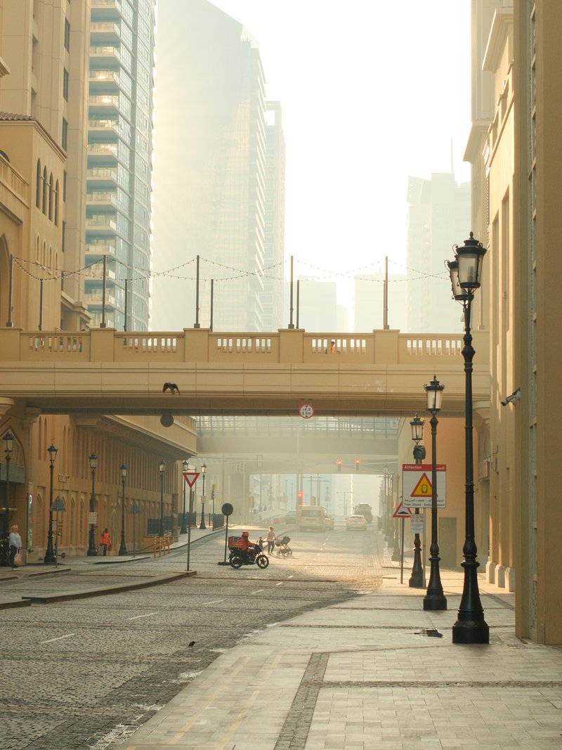 A quiet urban street with a bridge overhead, soft light filtering through buildings, creating a hazy atmosphere.