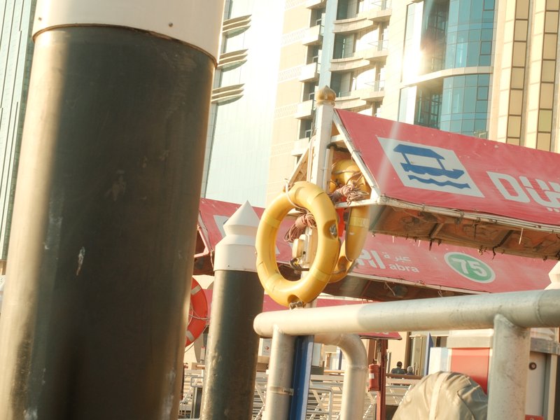 A yellow life ring hanging from a dock structure with buildings in the background under warm sunlight.