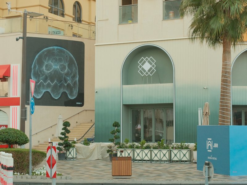 A modern urban scene featuring a large jellyfish mural on a building, with palm trees and a shopping area.