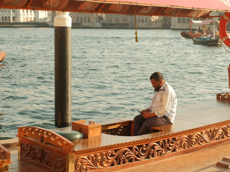A man sitting on a traditional wooden boat by the water, with city buildings in the background.