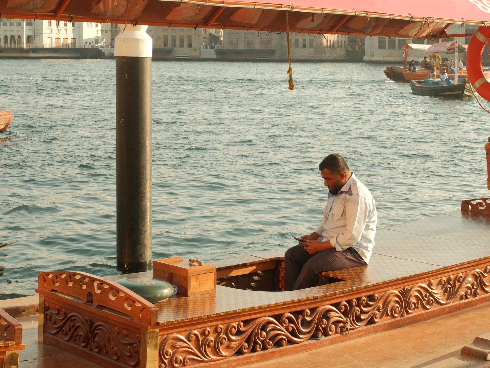 A man sitting on a traditional wooden boat by the water, with city buildings in the background.