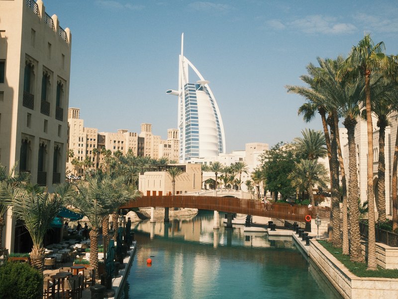 A view of the Burj Al Arab hotel with a bridge over a canal, surrounded by palm trees and buildings.