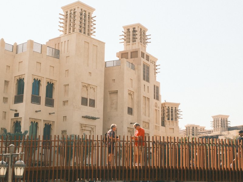 Two people standing on a bridge in front of traditional Middle Eastern architecture under bright sunlight.