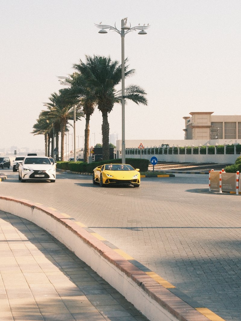 A yellow sports car drives along a palm-lined street with a white car in the background.