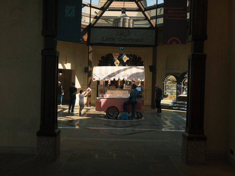 A vibrant indoor market scene with people interacting around a pink cart under a skylight.