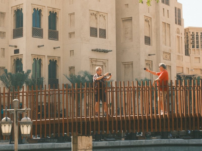 Two people on a wooden bridge in front of a building with palm trees, enjoying a sunny day.