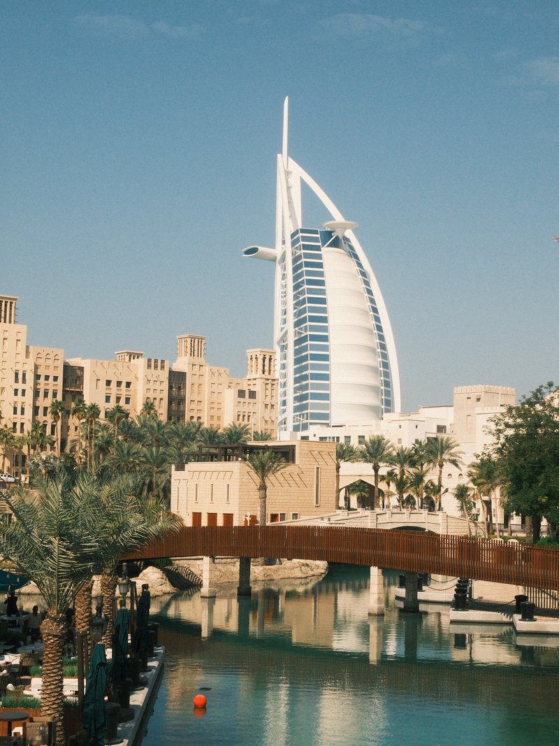 A modern skyscraper towers over traditional buildings beside a calm waterway with palm trees.