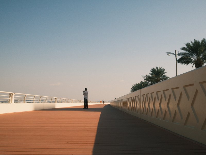 A lone figure standing on a wooden walkway beside a beach, with palm trees and a clear blue sky.