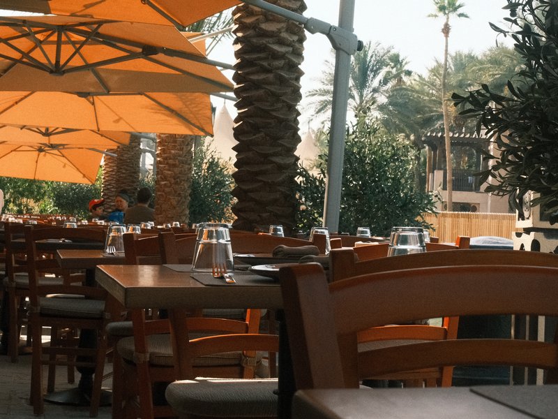 Outdoor restaurant scene with wooden tables, orange umbrellas, and palm trees in the background.