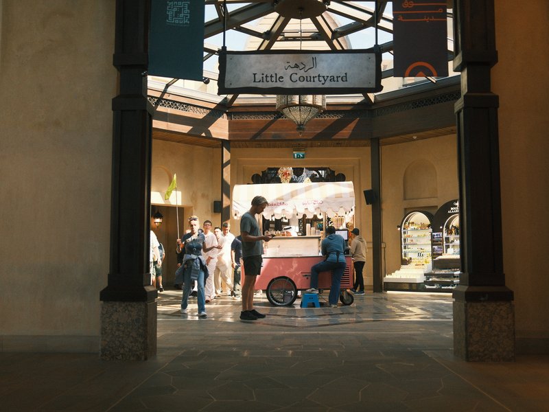 A bustling indoor market scene with people gathered around a food cart under a skylight.