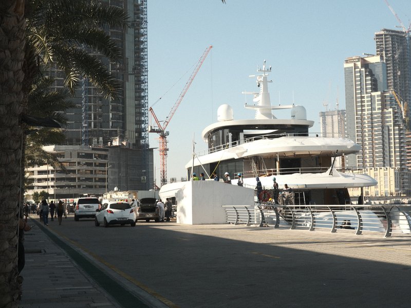 A luxury yacht docked near modern skyscrapers with palm trees lining the walkway.
