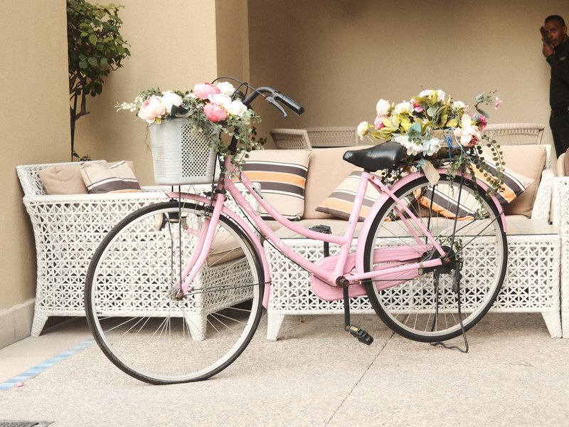 A pink bicycle parked beside a white wicker sofa adorned with flowers.