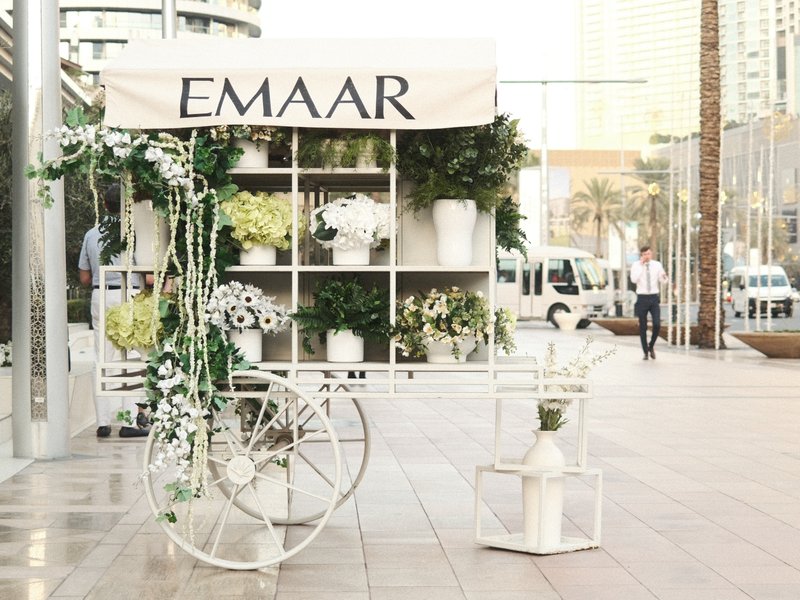 A floral display cart with the sign 'EMAAR' in an urban setting, surrounded by greenery and modern architecture.