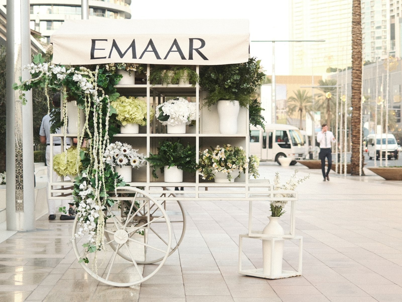 A floral display cart with the sign 'EMAAR' in an urban setting, surrounded by greenery and modern architecture.