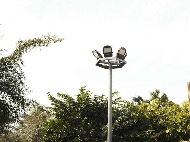 A tall light pole with multiple floodlights against a clear sky and green foliage.