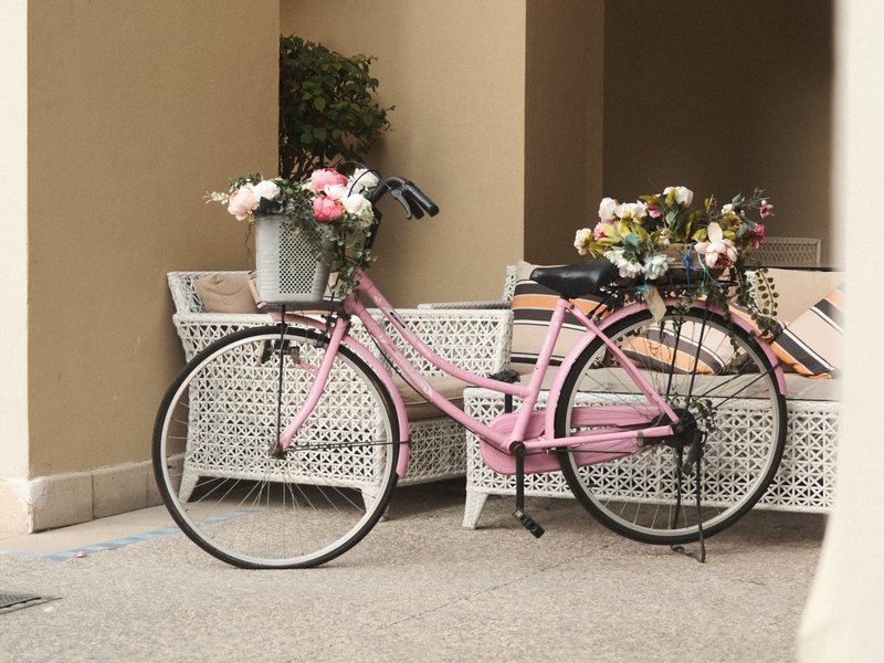 A pink bicycle with flower baskets parked beside a white wicker sofa in a cozy setting.
