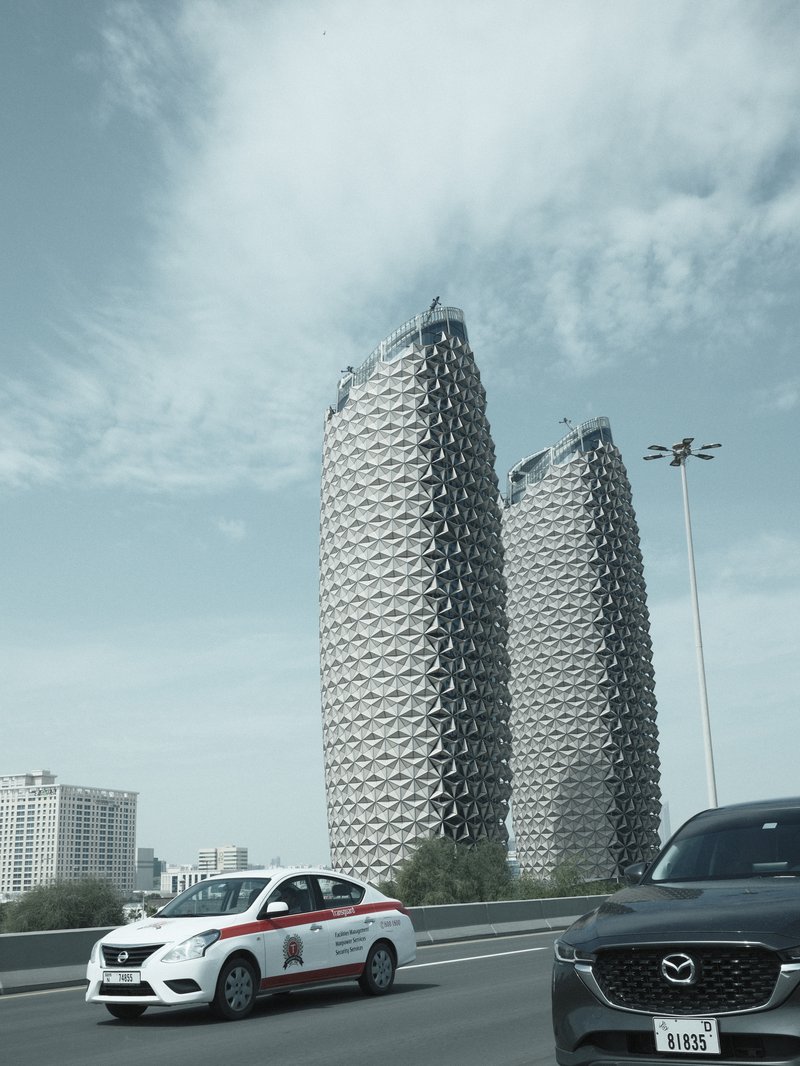 Two modern skyscrapers with unique architectural design under a cloudy sky, with cars in the foreground.