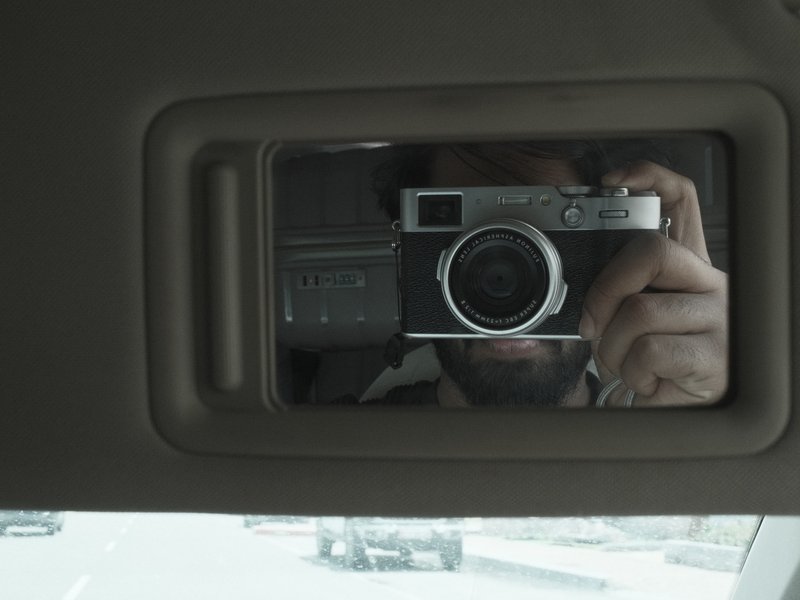 A person taking a selfie in a car's rearview mirror, capturing their camera and the road ahead.